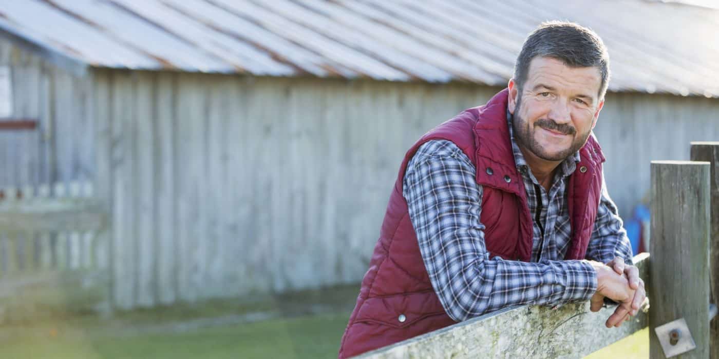 A farmer or rancher, mature man in his 50s, standing outside a barn, with his arms leaning on a wooden fence. He is wearing jeans, boots, a plaid shirt and vest.