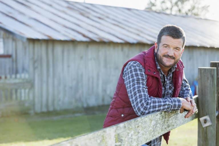 A farmer or rancher, mature man in his 50s, standing outside a barn, with his arms leaning on a wooden fence. He is wearing jeans, boots, a plaid shirt and vest.