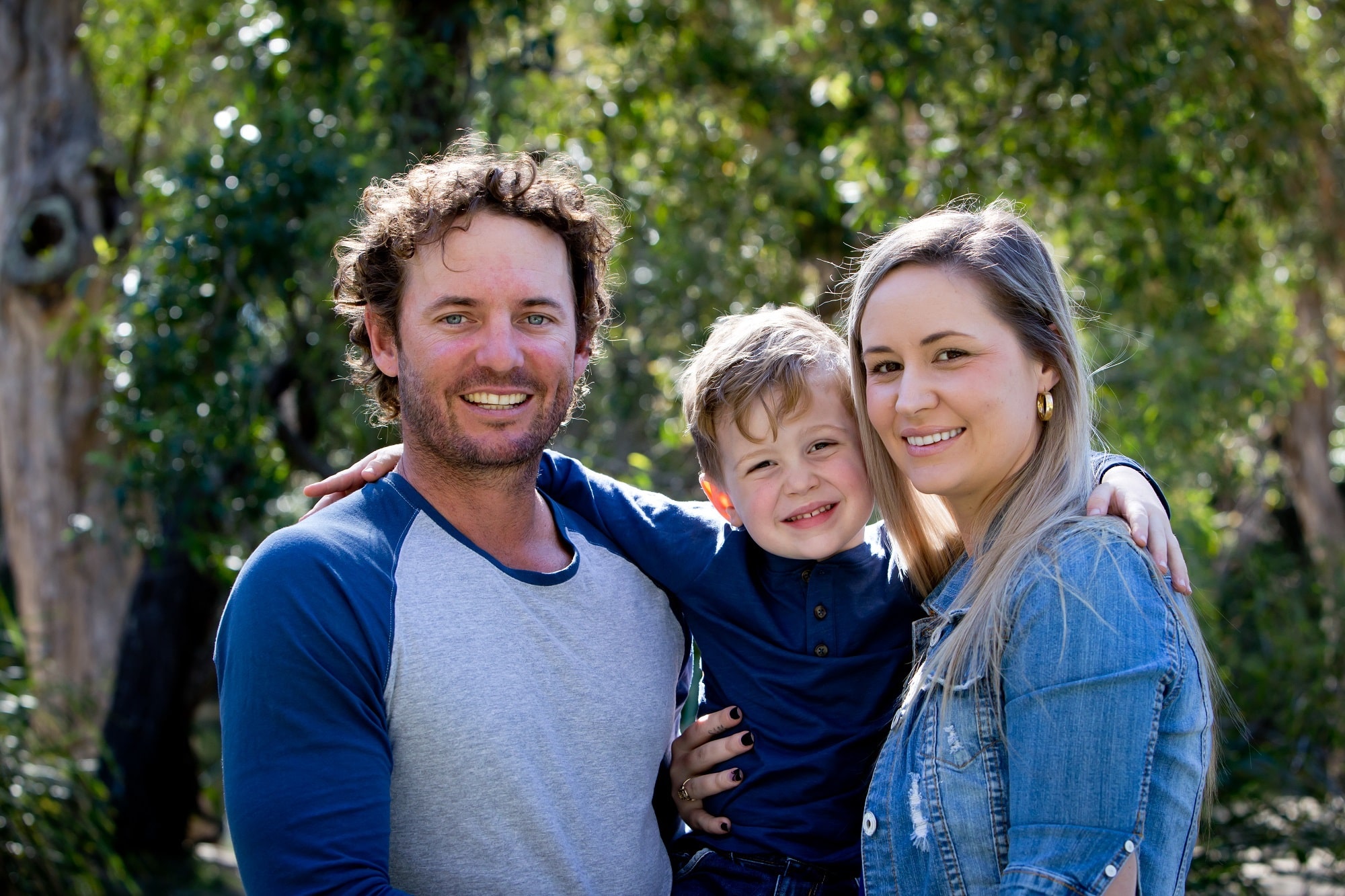 Father, Mother and young son together outdoors in sunlight in park wearing casual clothing - looking at camera