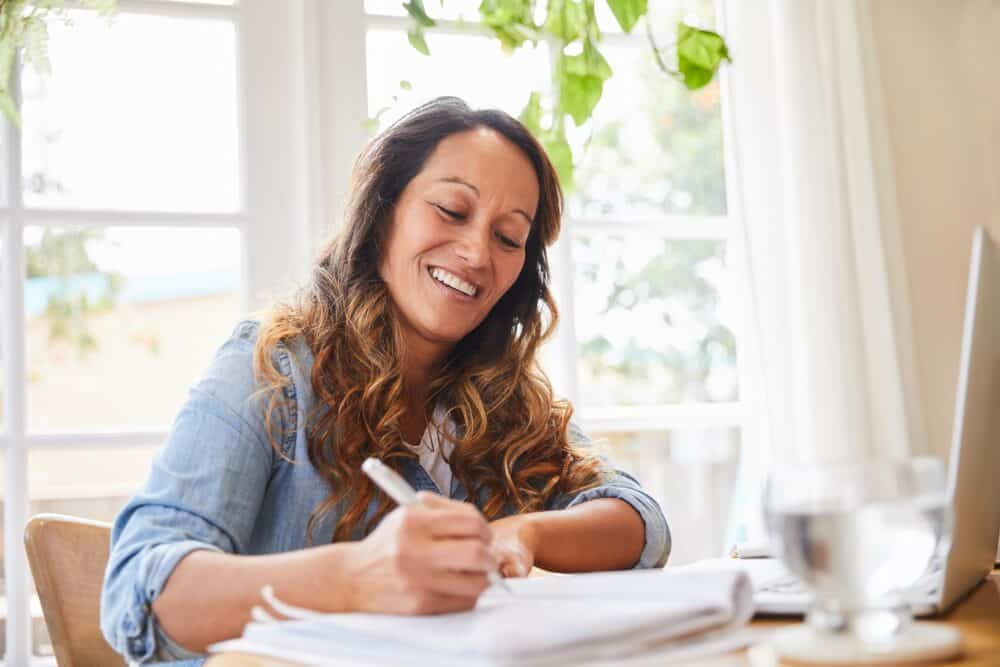 Smiling mature woman writing down ideas in a notebook while working at a table in her living room at home