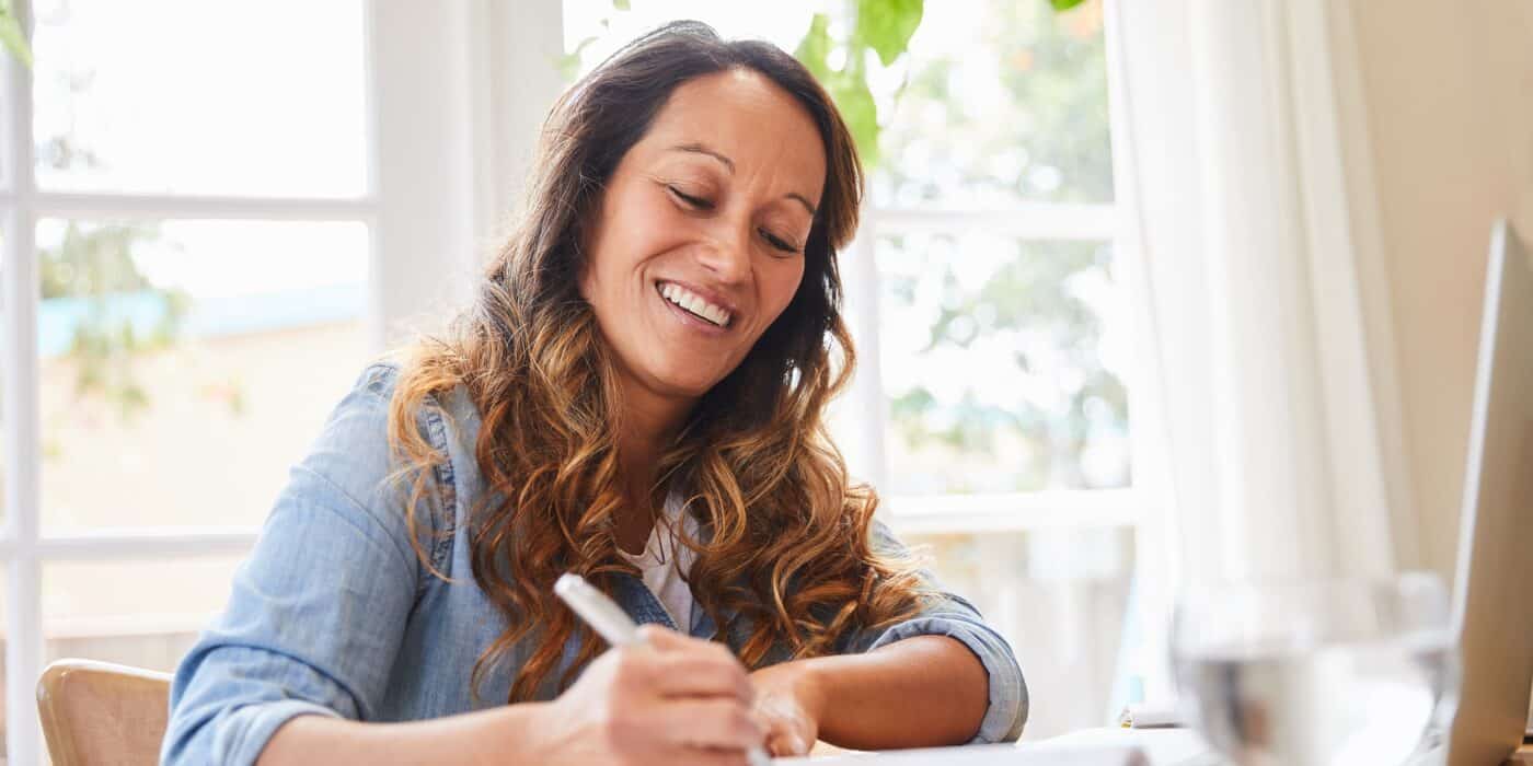 Smiling mature woman writing down ideas in a notebook while working at a table in her living room at home