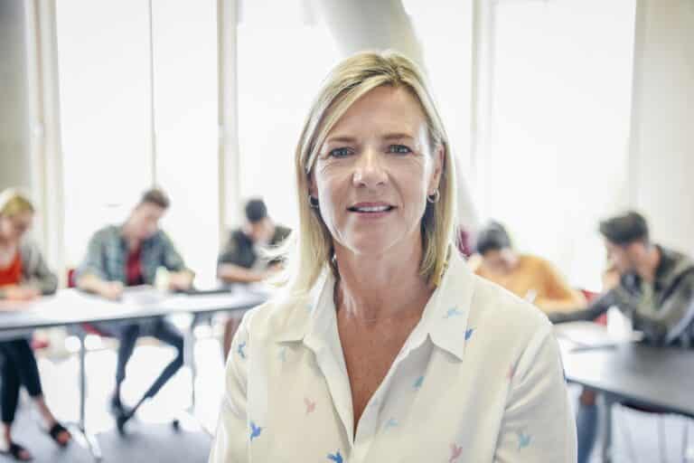 Female lecturer in classroom with contented expression, college students writing at desks behind her