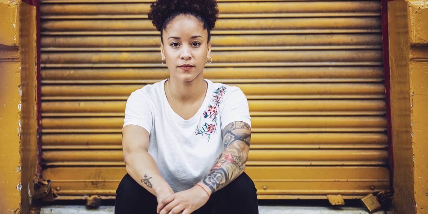 Portrait of woman sitting against closed yellow shutter. Confident female is having tattoo on hands. She is in fashionable casuals.