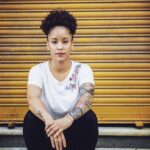 Portrait of woman sitting against closed yellow shutter. Confident female is having tattoo on hands. She is in fashionable casuals.