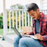 Man sitting outside a house working on laptop and writing notes.