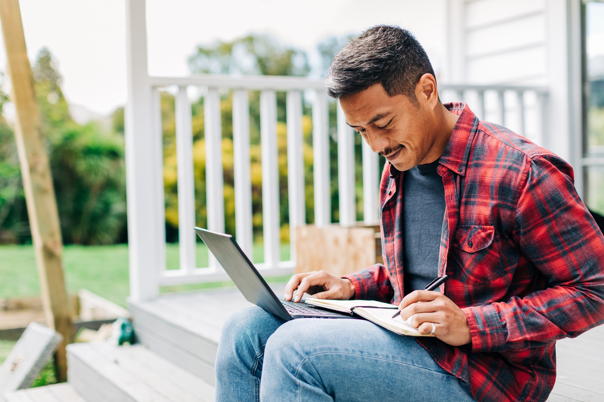 Man sitting outside a house working on laptop and writing notes.