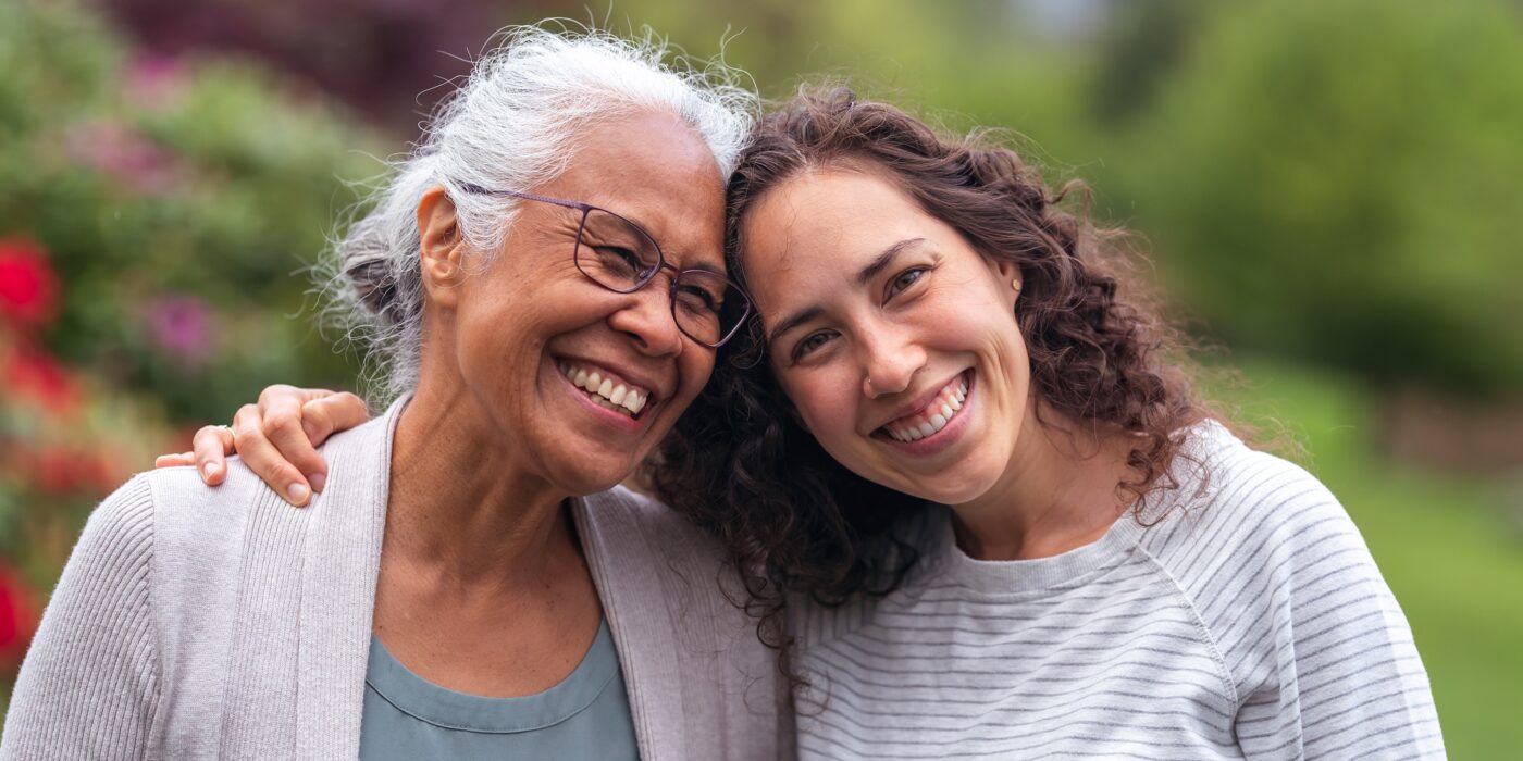 A senior woman embraces her millennial daughter as they happily walk through a natural parkland area and enjoy their time together. The daughter is smiling at the camera.